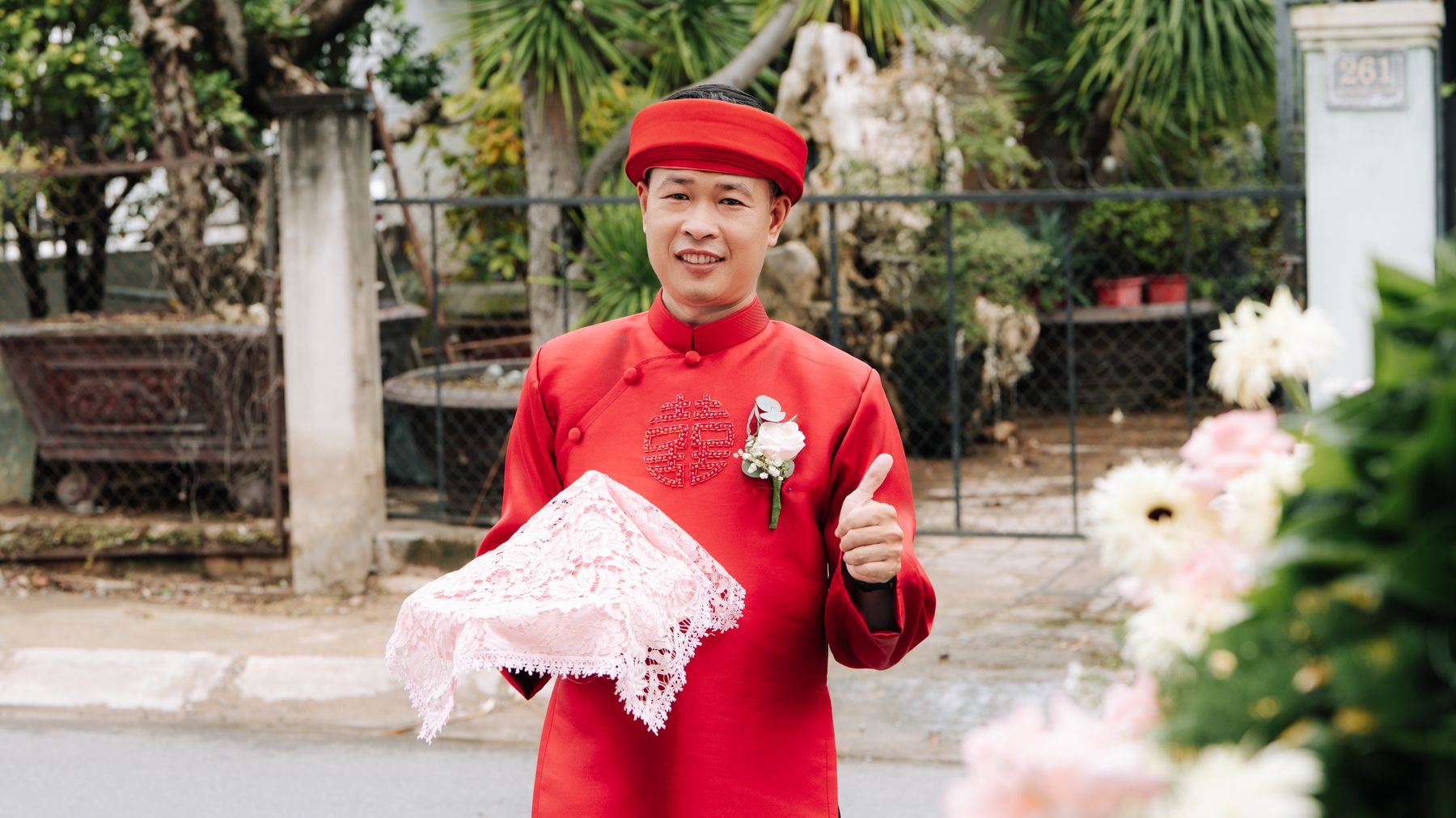 Man in traditional Vietnamese red wedding attire holding a gift box outdoors, smiling.