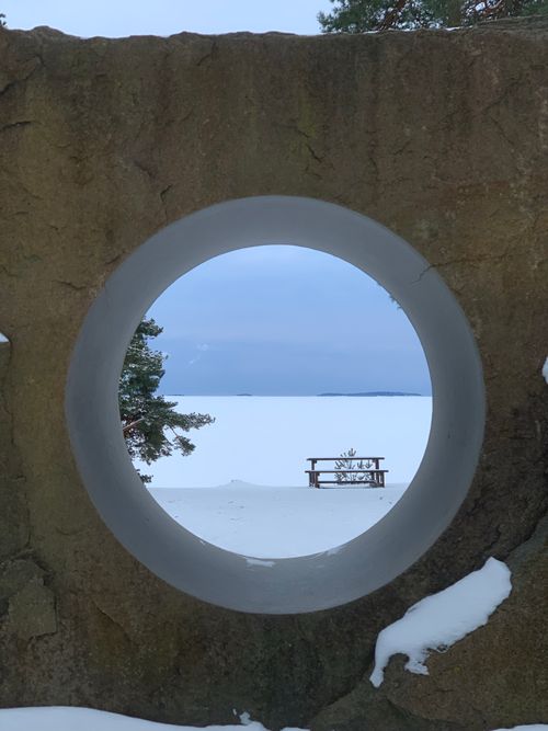 Free A serene winter scene viewed through a circular stone frame, featuring a snowy landscape with a lone picnic table. Stock Photo