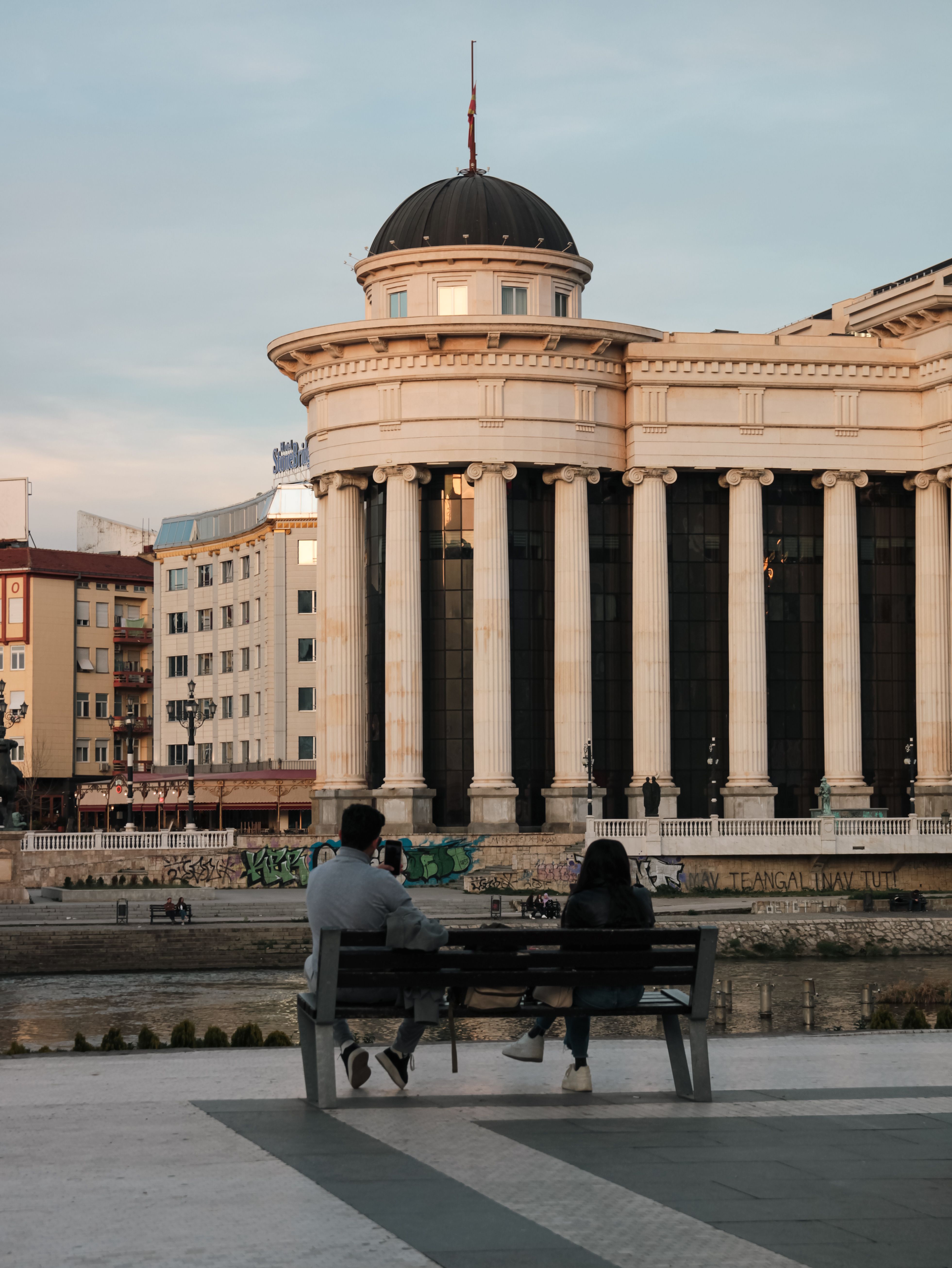 Two people seated on a bench near the Archaeological Museum in Skopje, North Macedonia.