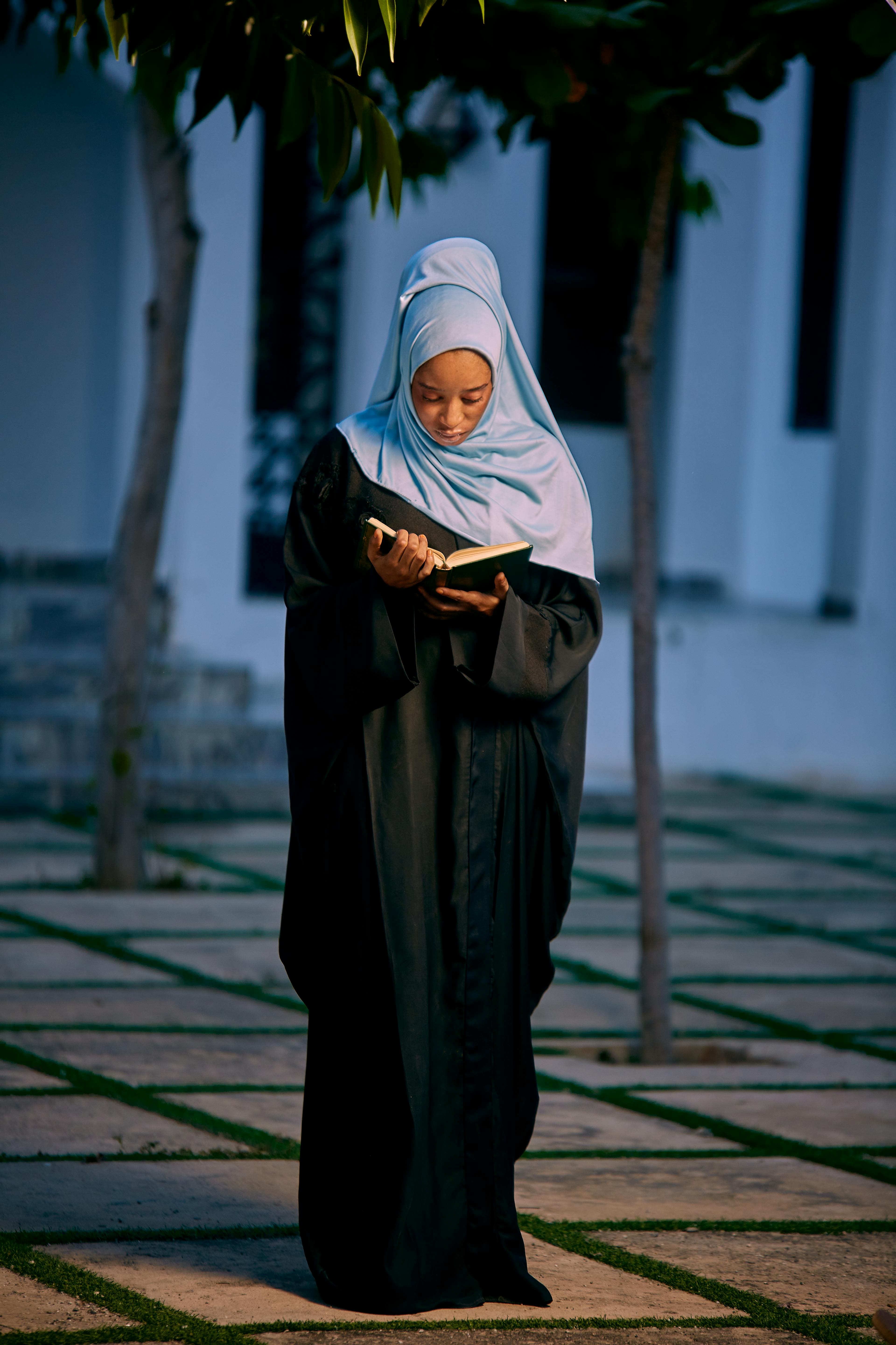 A woman in a hijab reads a book outdoors at twilight, conveying peace and contemplation.