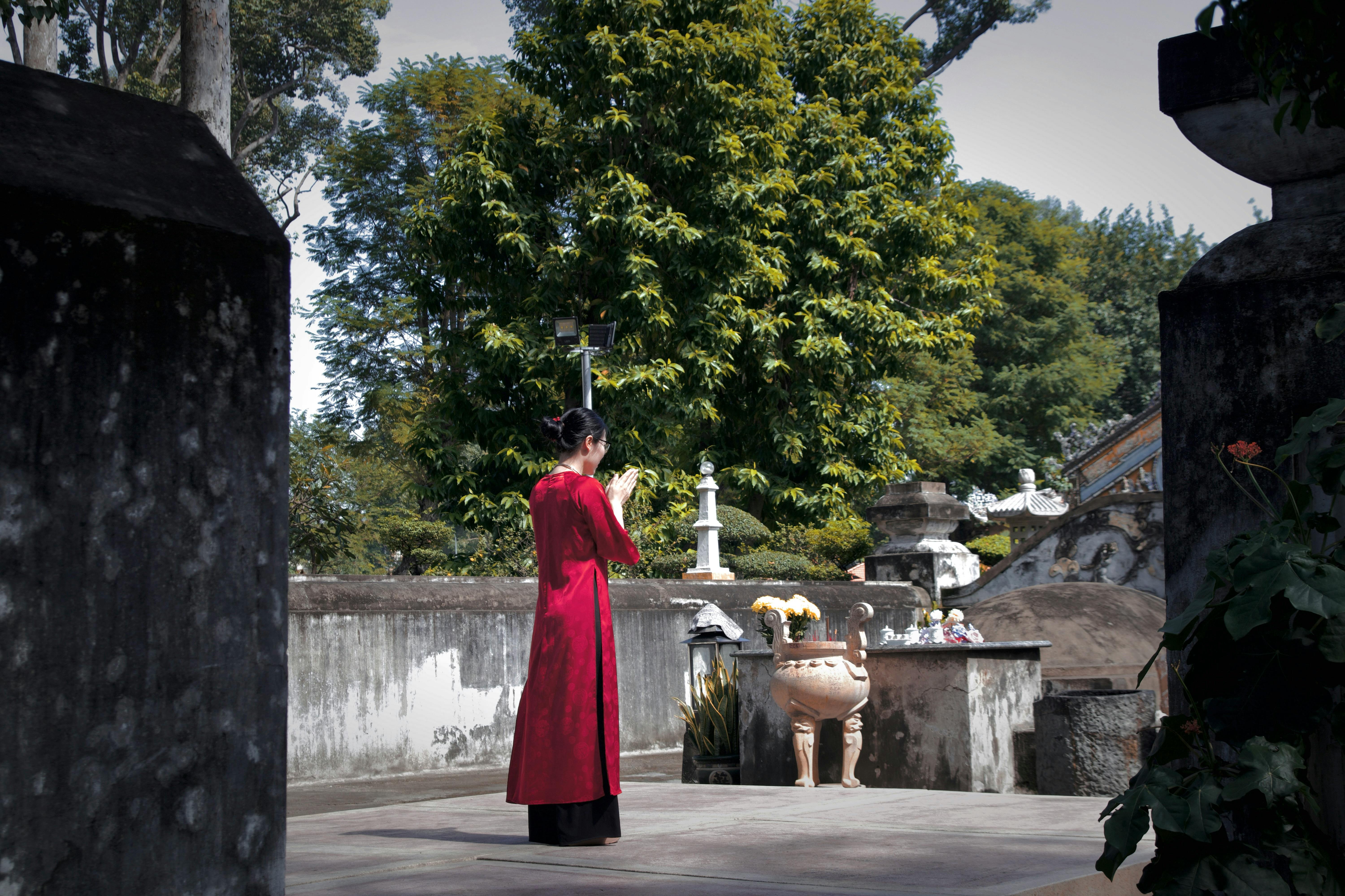 An Asian woman in a red dress prays at an outdoor shrine surrounded by greenery.