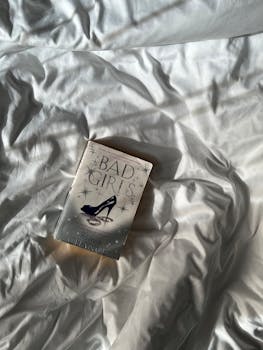 A book titled 'Bad Girls' rests on white crumpled bedding under soft morning sunlight.