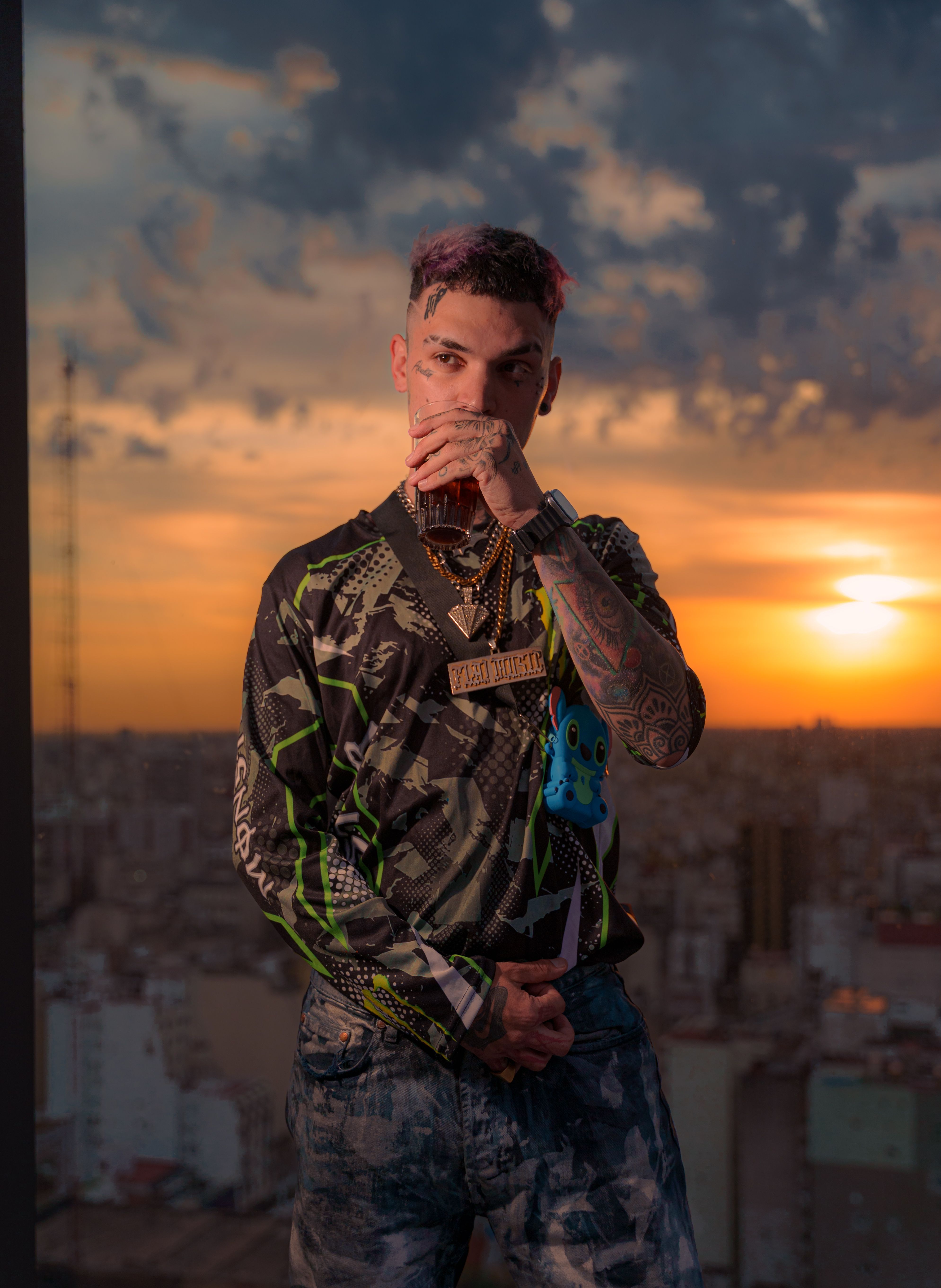 A tattooed man in urban attire poses against a vibrant sunset cityscape.
