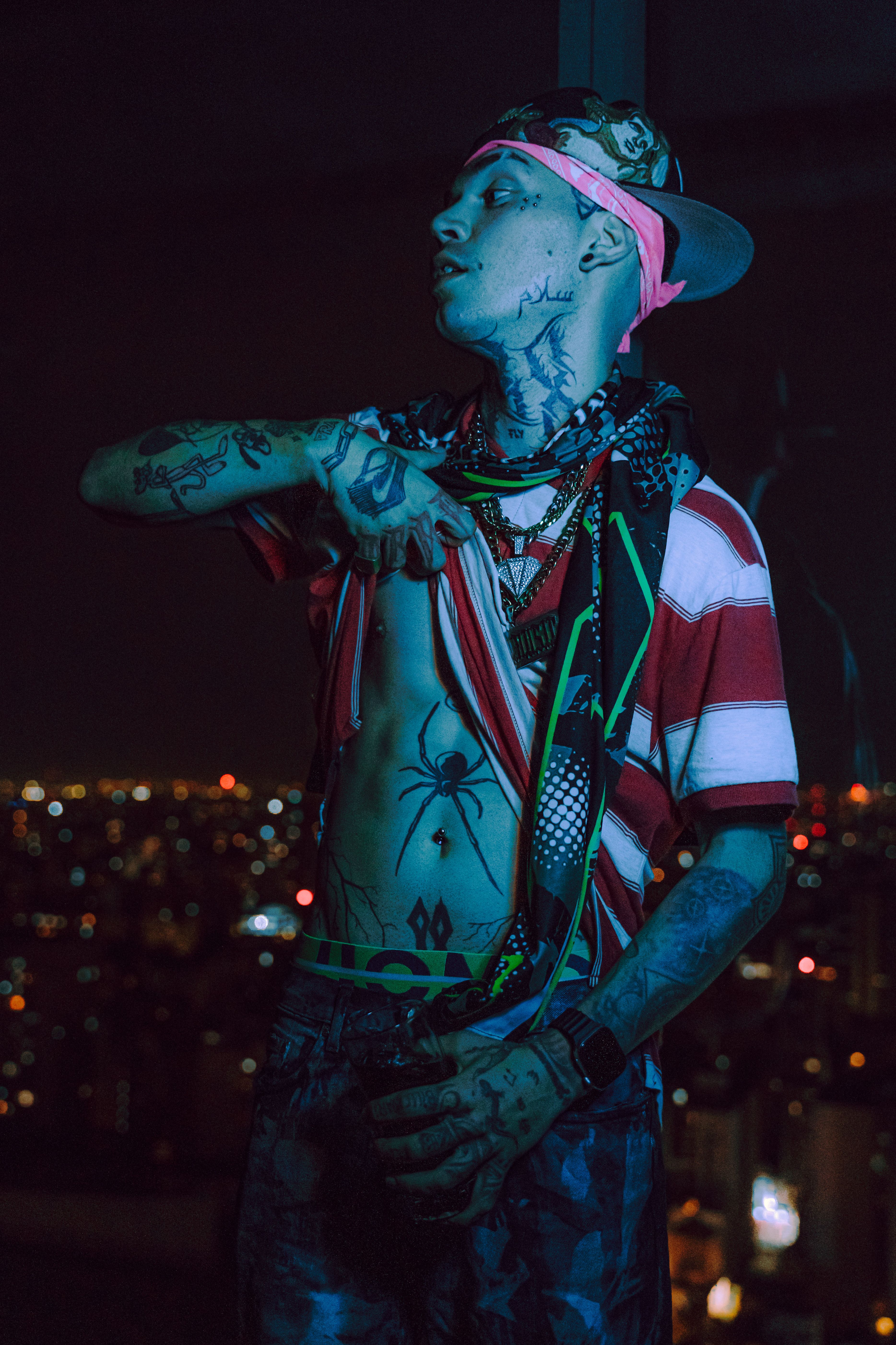 Stylish tattooed man poses confidently with vibrant city lights in the background.