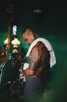 Tattooed man with towel stands in a stylish barbershop, reflecting modern fashion and lifestyle.