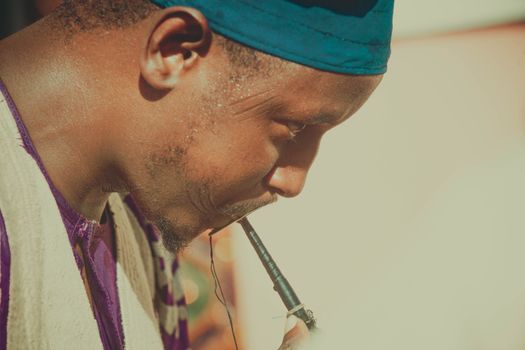 African musician playing a traditional pipe instrument outdoors under bright sunlight.
