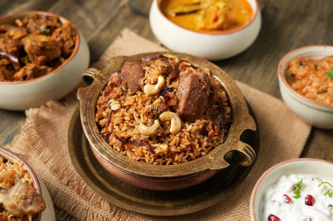 Meat biryani with cashews in a brass pot surrounded by curry and raita bowls