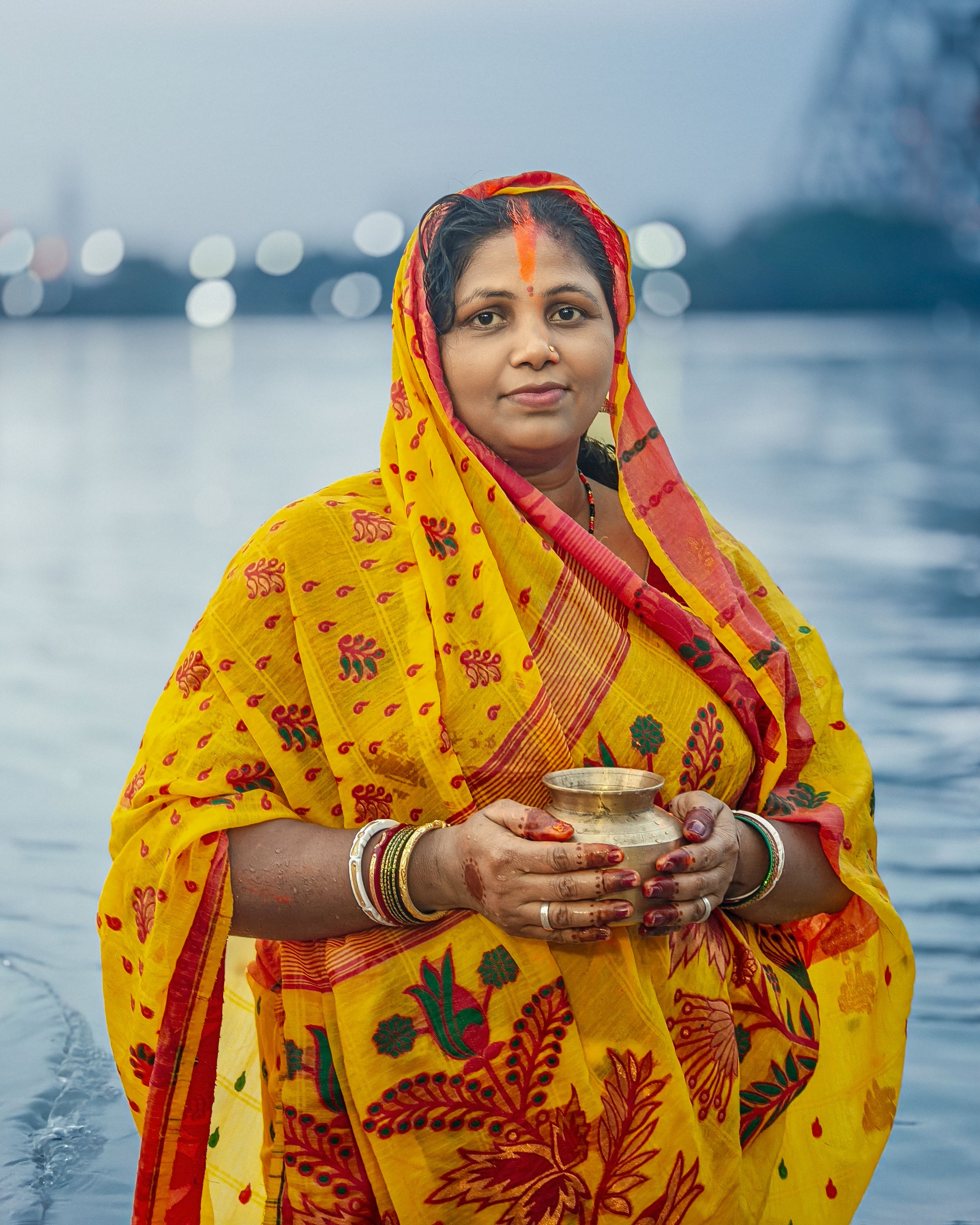 South Asian woman wearing a vibrant saree holding a pot by the river during a ritual.
