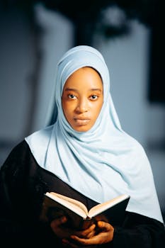 Portrait of a young woman wearing a hijab, holding an open book, with a focused expression.