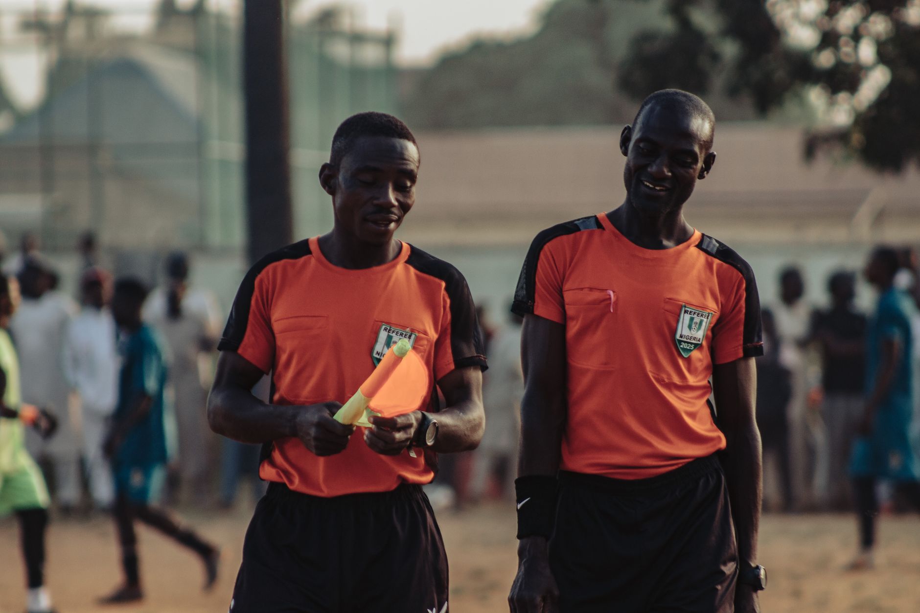 Two soccer referees in orange jerseys walking during a daytime match.