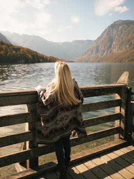 Woman with long blonde hair stands on a balcony overlooking a tranquil mountain lake.