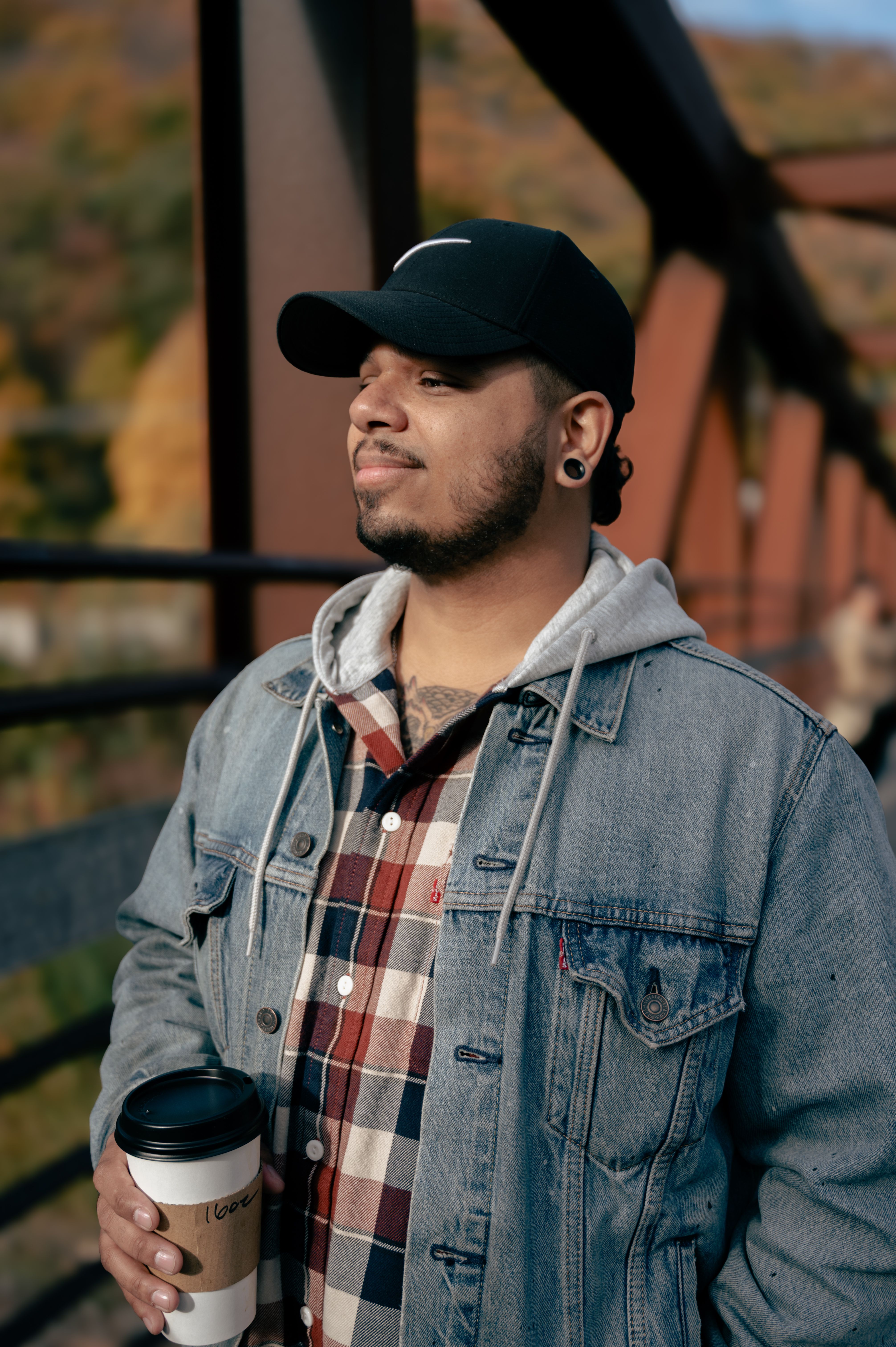 Man in denim jacket holding coffee on an autumn bridge walk.