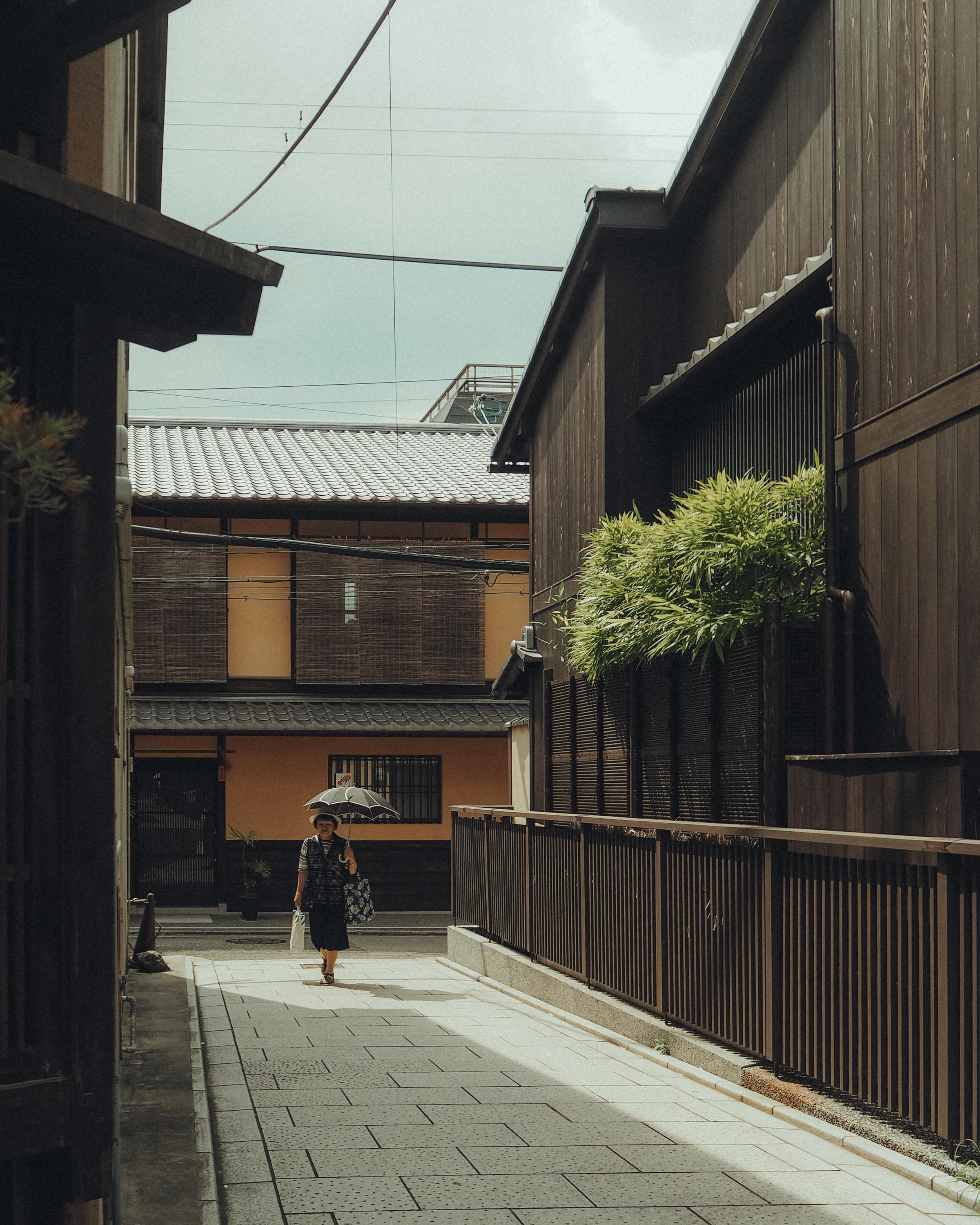 A woman with an umbrella walks down a narrow street in a traditional Japanese neighborhood, framed by wooden houses.