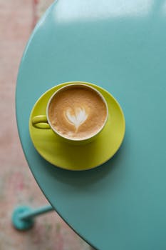 Top view of a latte with heart-shaped art in a yellow cup on a teal table.