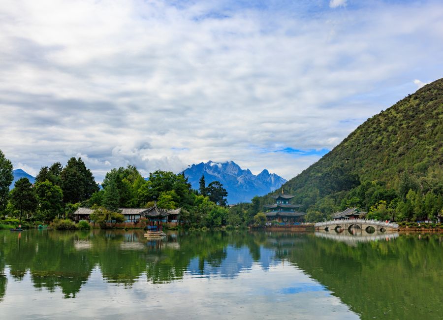 Peaceful scene of a scenic park with mountains, lake, and traditional Chinese architecture under a cloudy sky.