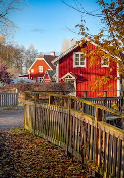A traditional red wooden house surrounded by autumn foliage with a rustic fence in a Nordic countryside.