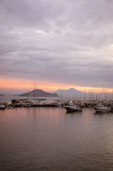 Peaceful harbor scene at twilight with boats, mountains, and a vivid sunset sky.