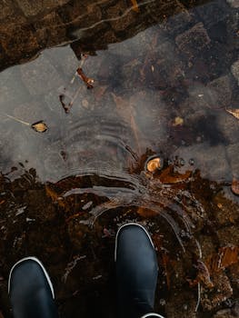 Black boots stand in a rain puddle with autumn leaves.