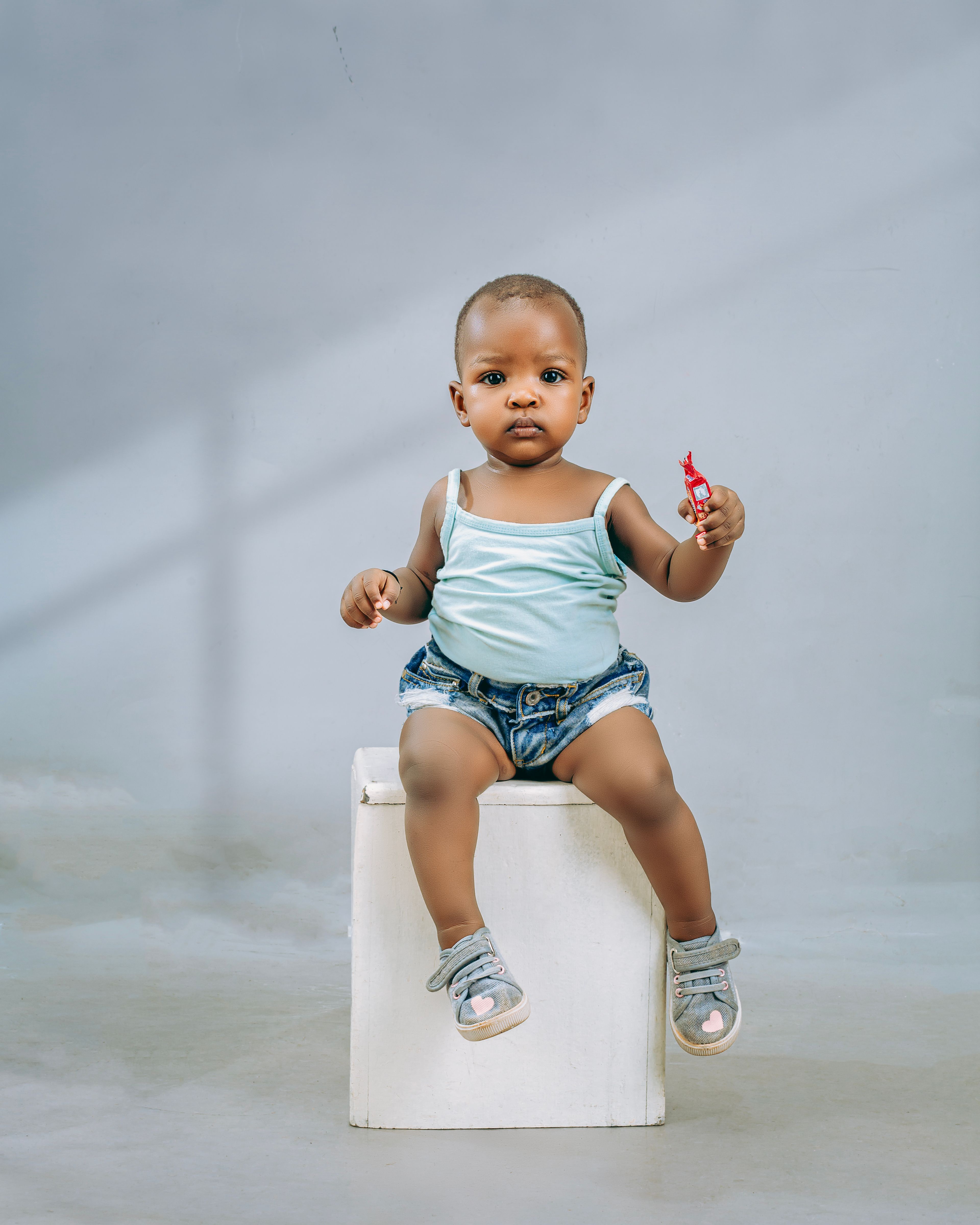 Portrait of a cute baby sitting on a white box indoors holding a red toy, wearing casual clothes.