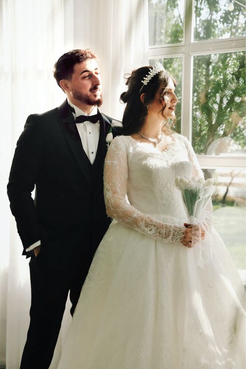 Free Bride and groom posing by a sunlit window, looking elegant and joyful. Stock Photo