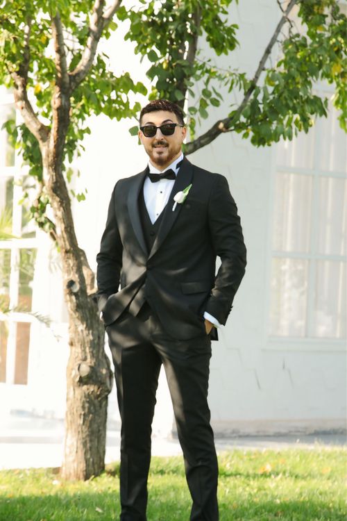 Free Groom in a black tuxedo posing outdoors under a tree, exuding elegance and style. Stock Photo