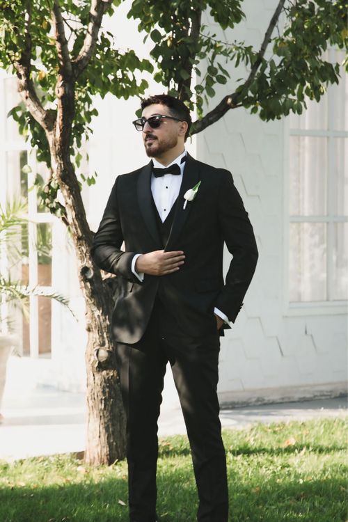 Free Stylish man in a black tuxedo standing confidently outdoors with greenery and sunlight. Stock Photo