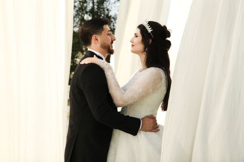 Free Elegant bride and groom embracing in wedding attire amidst flowing white curtains. Stock Photo