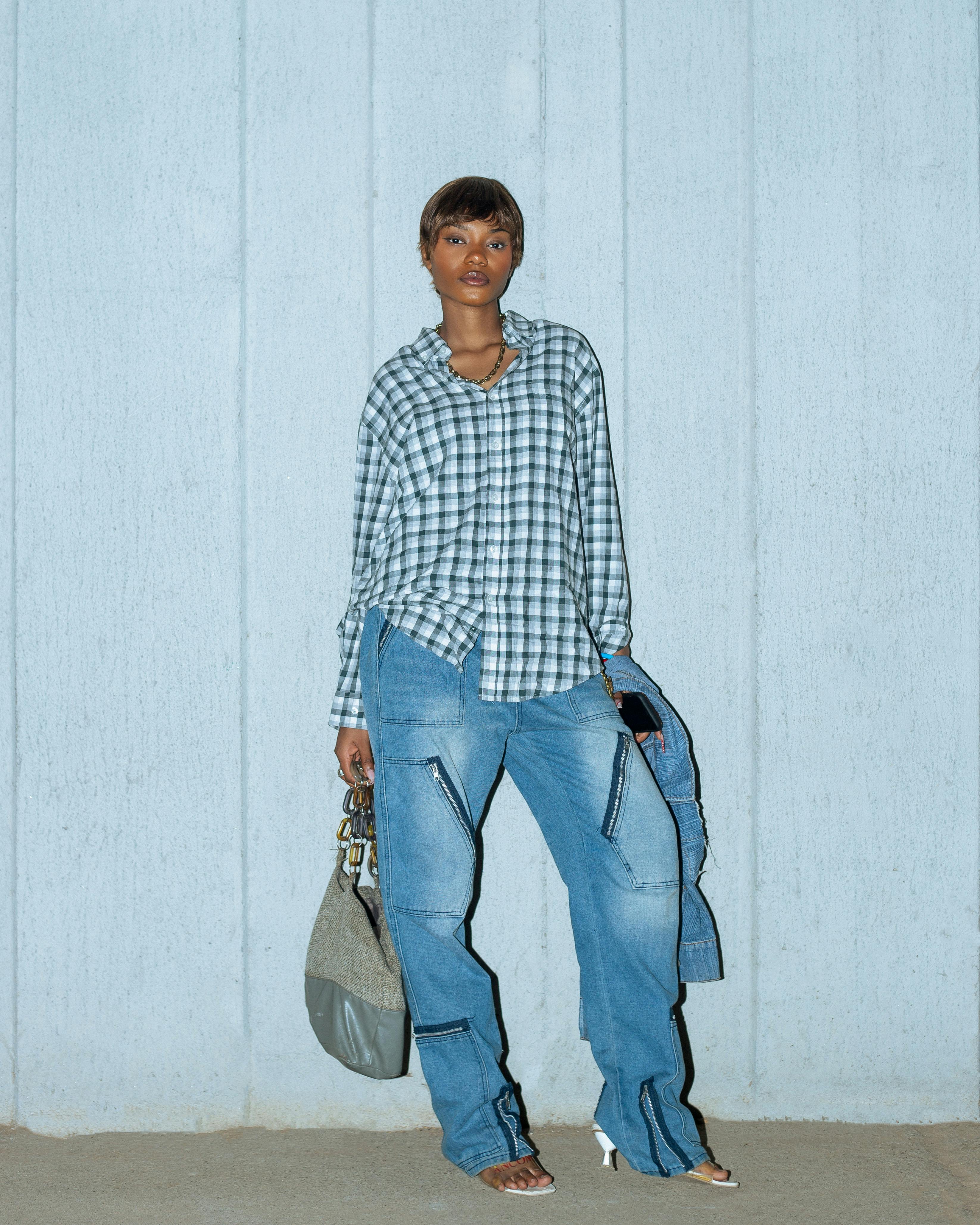 Gratis Moderigtig kvinde poserer i Nigeria iført en ternet skjorte og jeans op mod en tekstureret væg. Lagerfoto