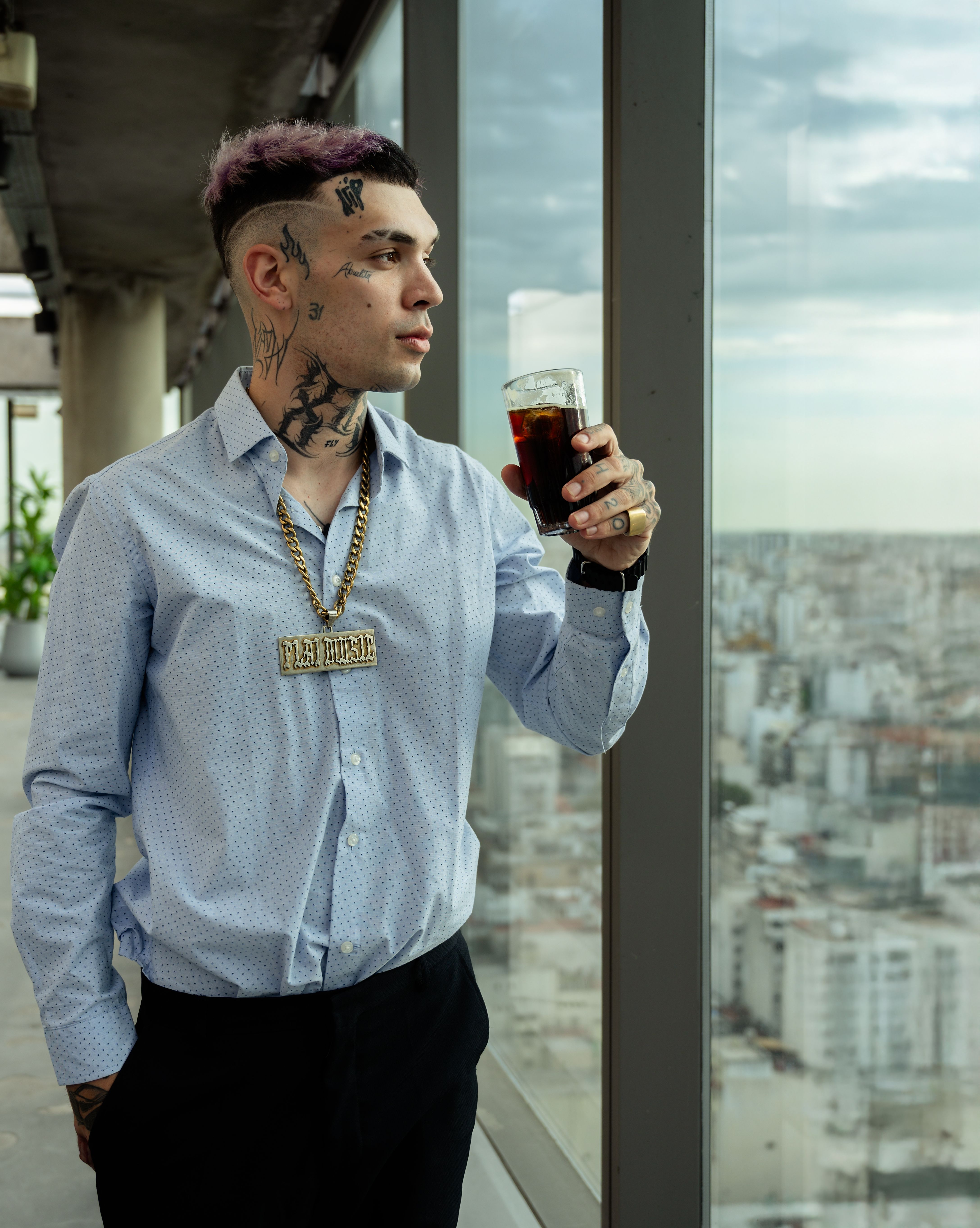 Young tattooed man in blue shirt holding a drink, gazing out over Buenos Aires city skyline.