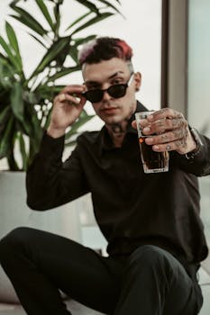 A fashionable tattooed man holds a drink in a modern indoor setting, exuding a cool and relaxed vibe.