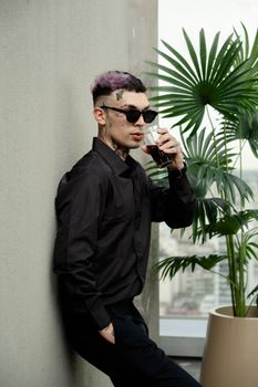 Trendy tattooed man in black attire drinks on a balcony with city view in Buenos Aires.