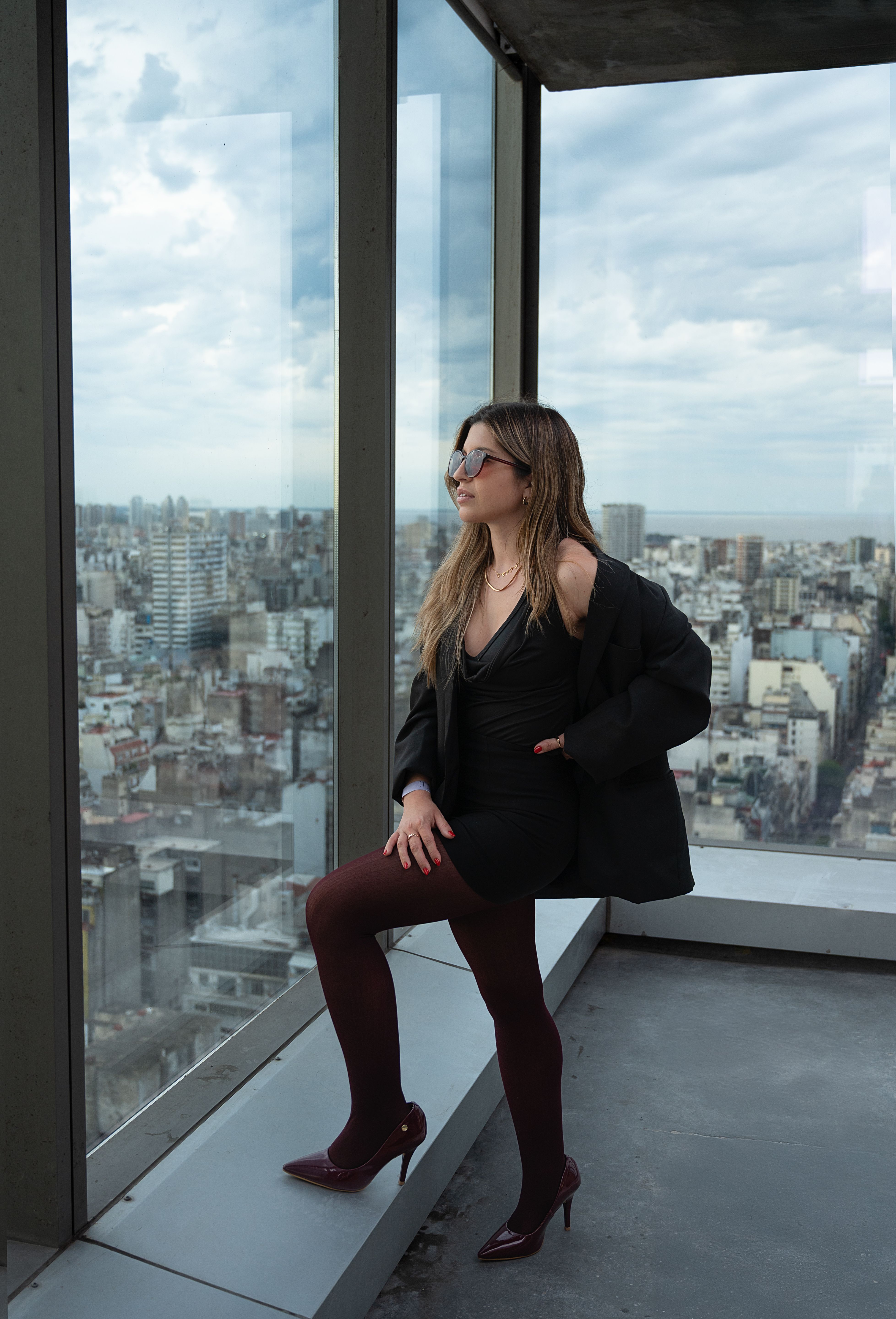 Fashionable woman in black dress and coat poses by a large window with a stunning view of Buenos Aires.