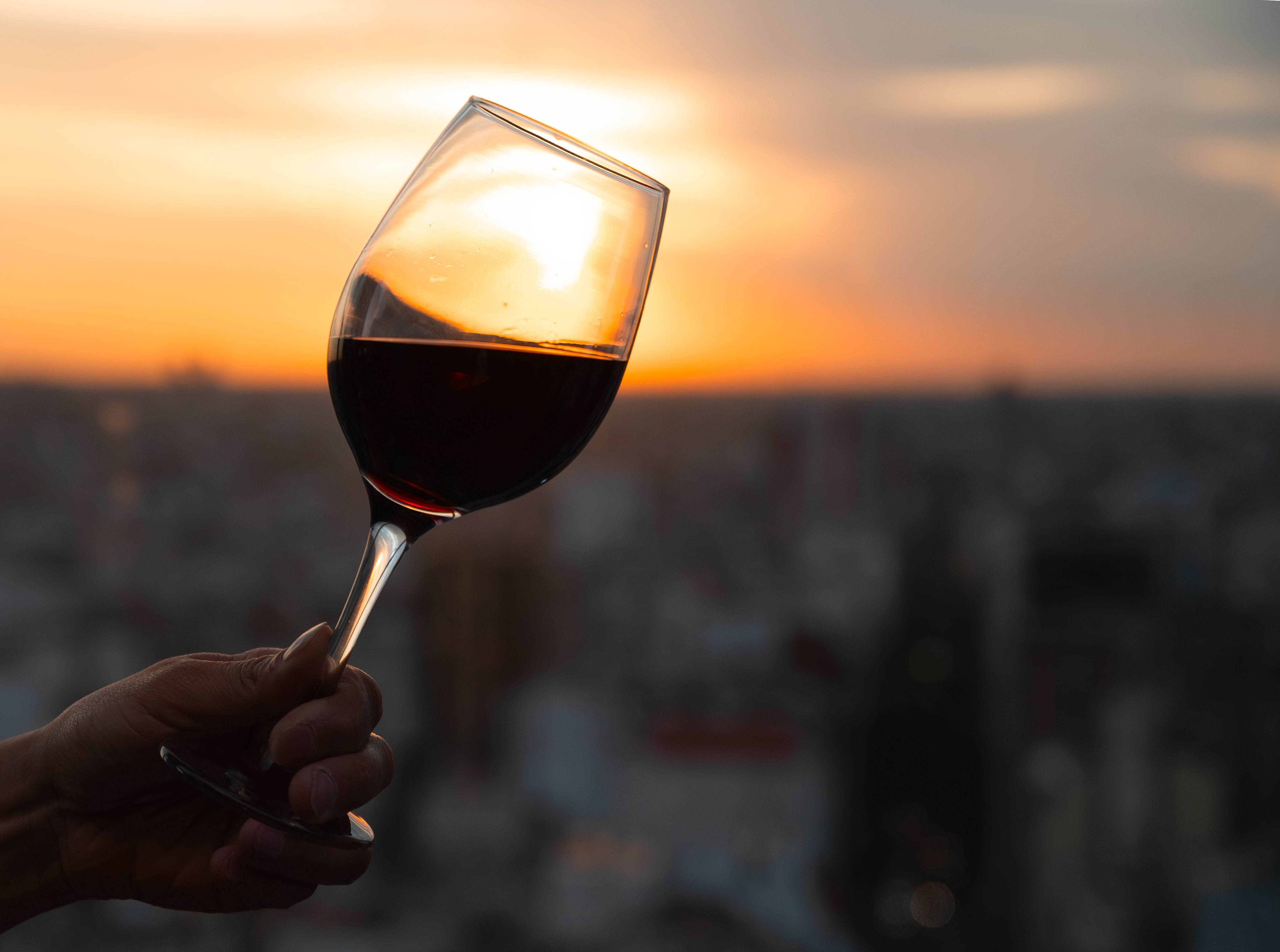 Hand holding a glass of red wine against a golden sunset overlooking a blurred city skyline