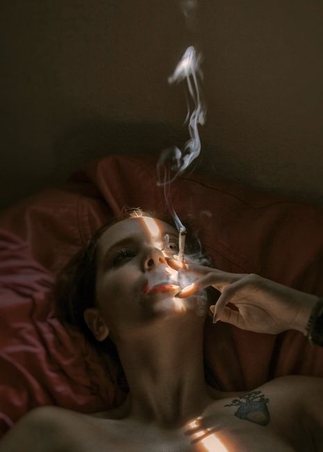 A woman lying on a bed indoors, smoking a cigarette with dramatic lighting casting shadows on her face.