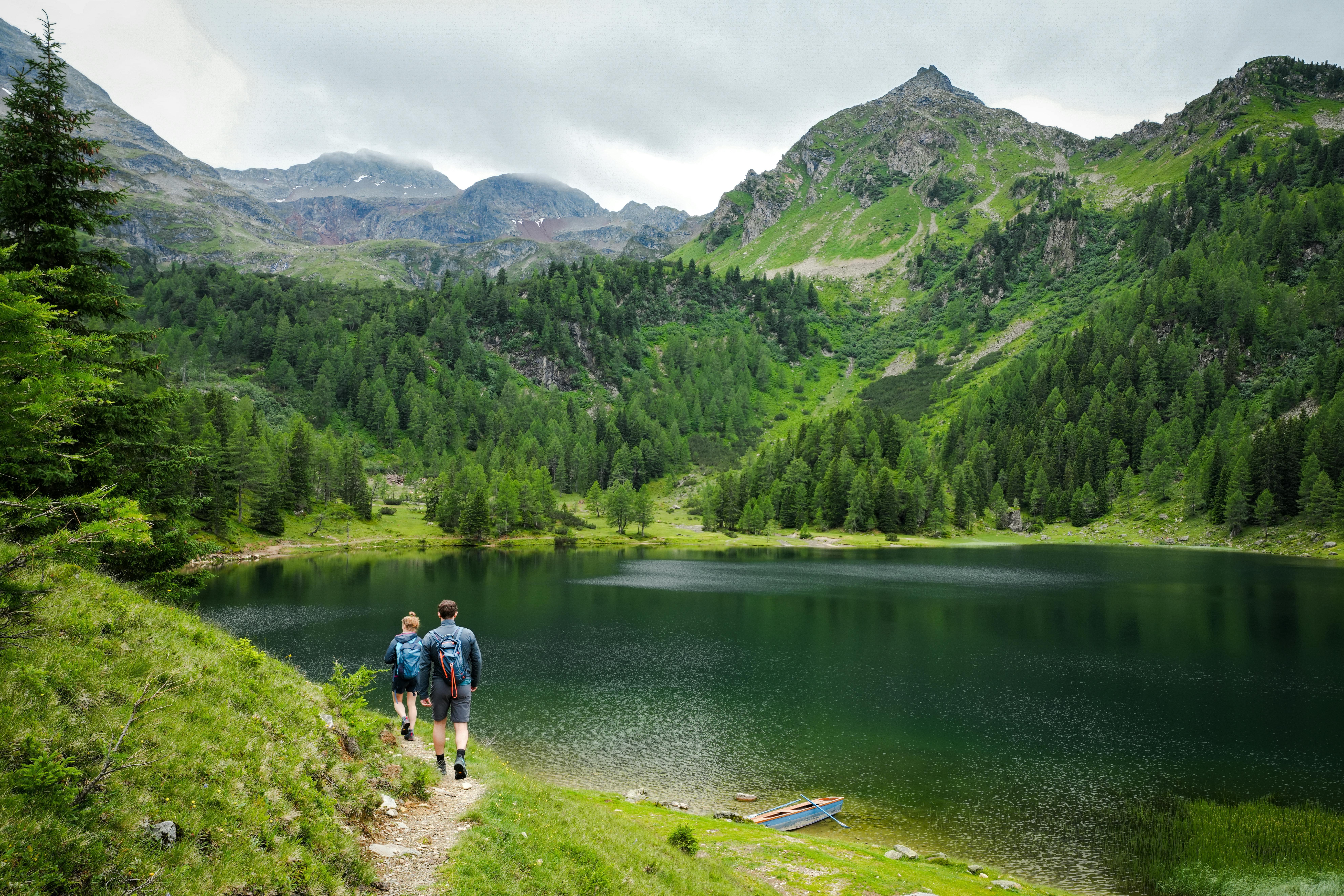 Two hikers enjoy a serene path by an alpine lake in Steiermark, Austria, surrounded by lush green mountains.