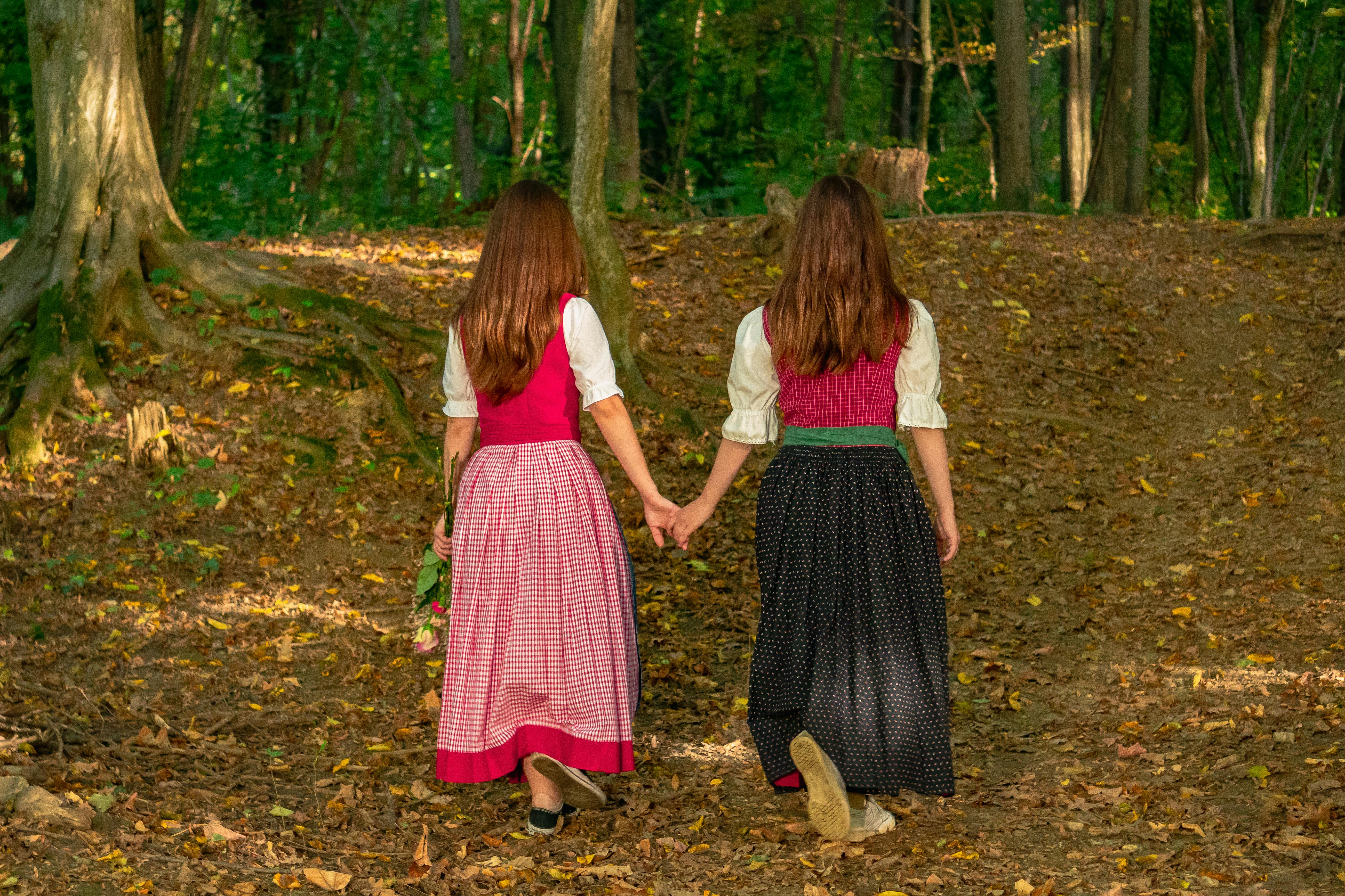 Two women holding hands and walking through a wooded area in traditional Austrian attire.