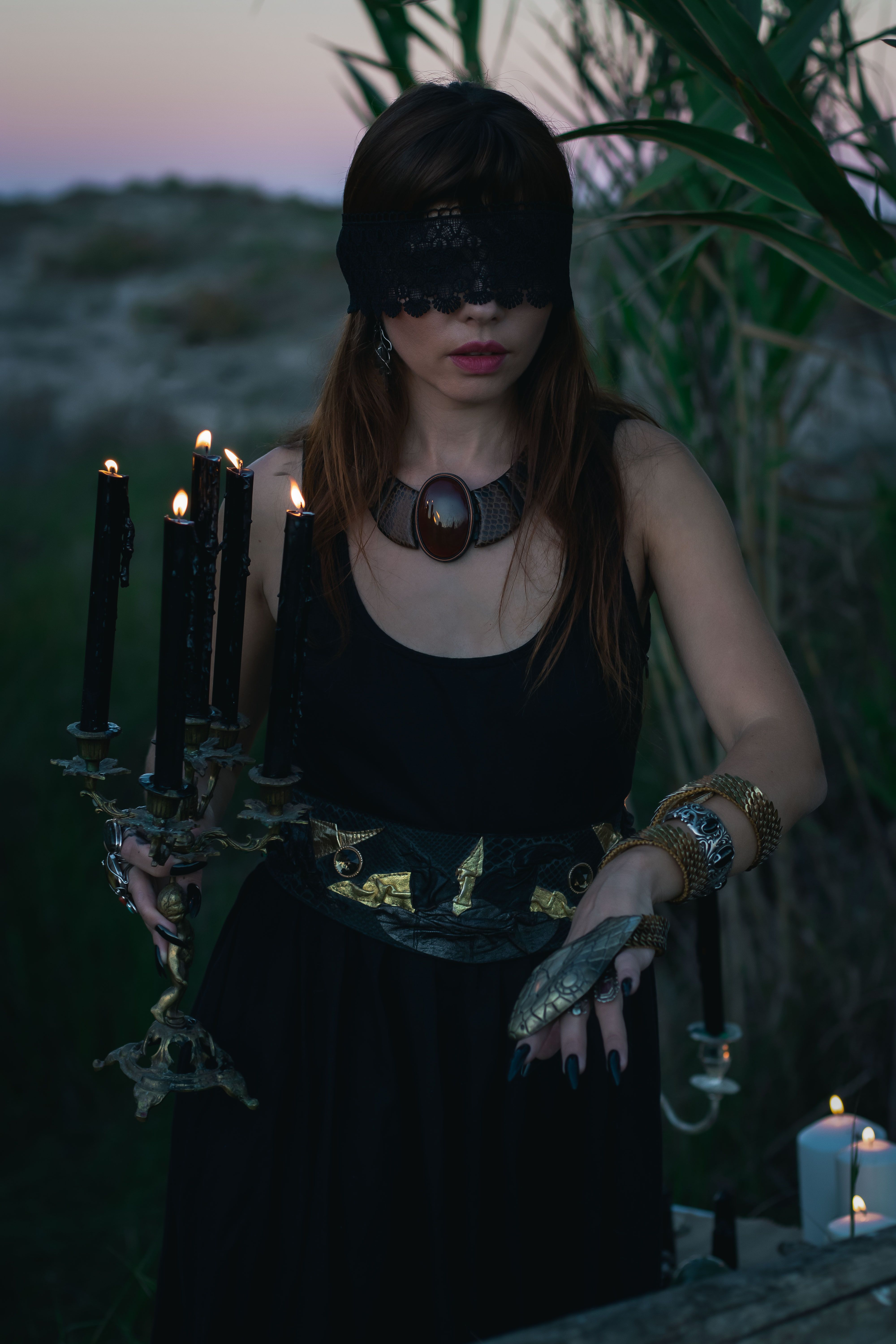 Woman in dark attire, blindfolded, holding a gothic candelabra outdoors at twilight.