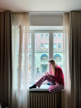 A young woman sitting on a window sill, using her phone in a Helsinki hotel room, Uusimaa, Finland.