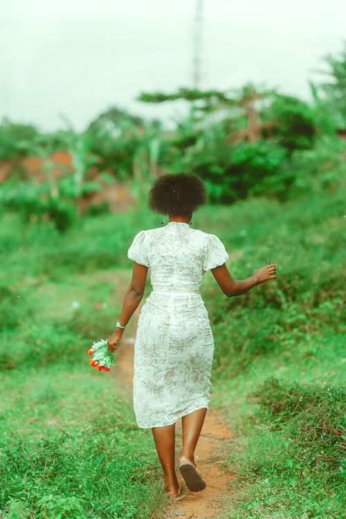 Free A woman in a lace dress holds flowers while walking along a lush green pathway outdoors. Stock Photo
