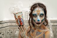 Woman with intricate Day of the Dead face paint holding decorated item indoors.