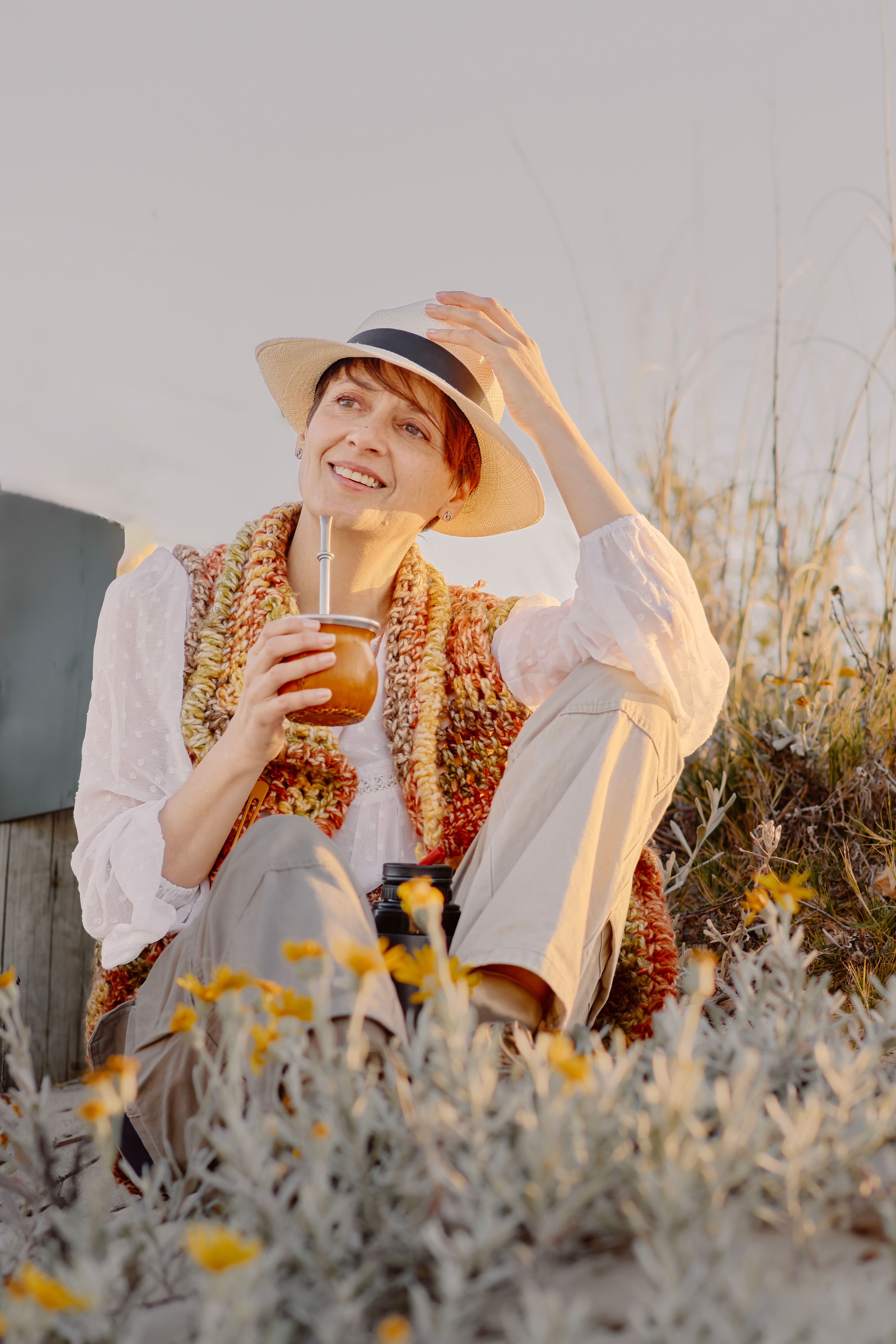 Smiling woman in hat enjoys a sunny day outdoors with a beverage.