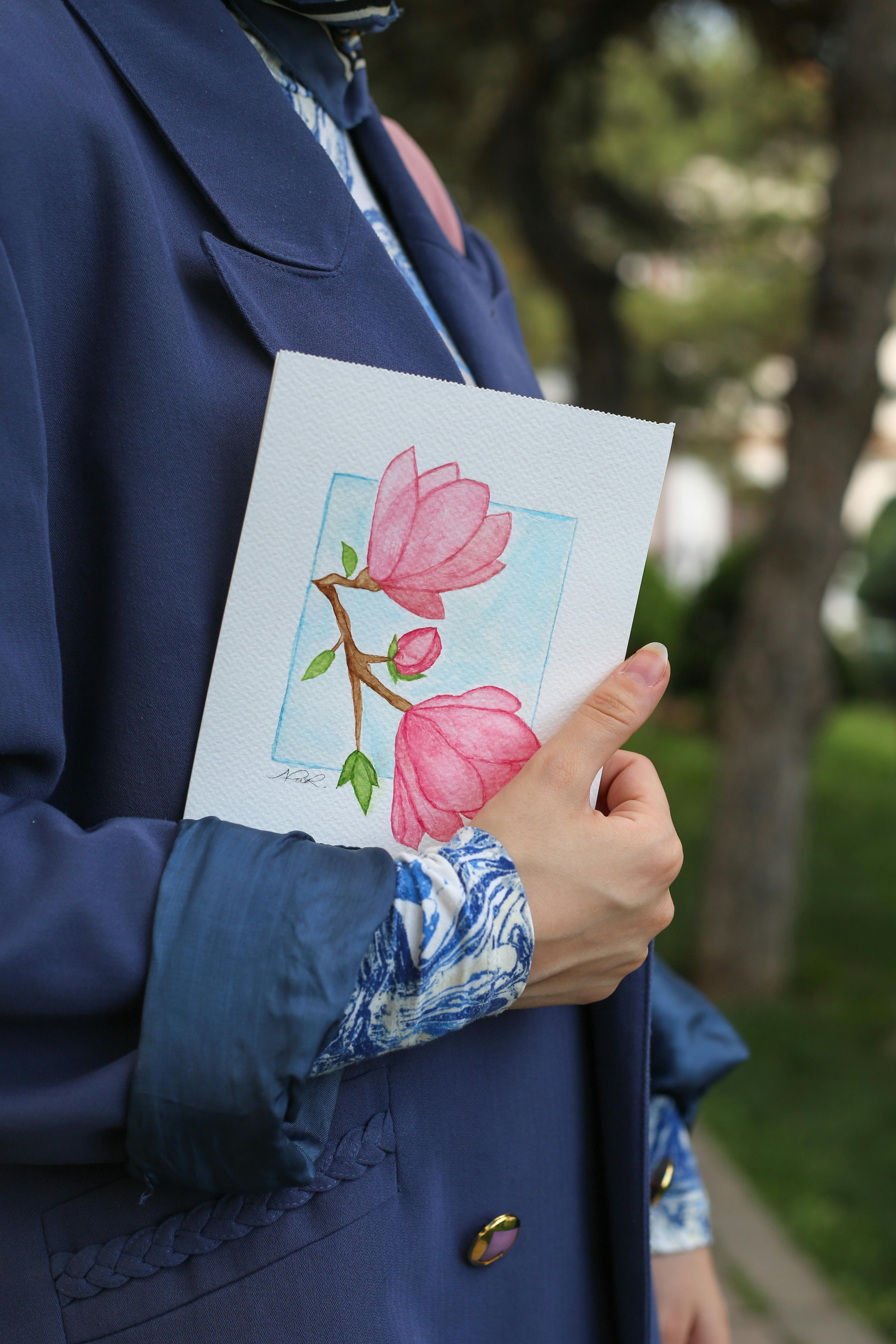 Woman in blue coat holds a floral watercolor painting while walking outdoors, adding an artistic touch.