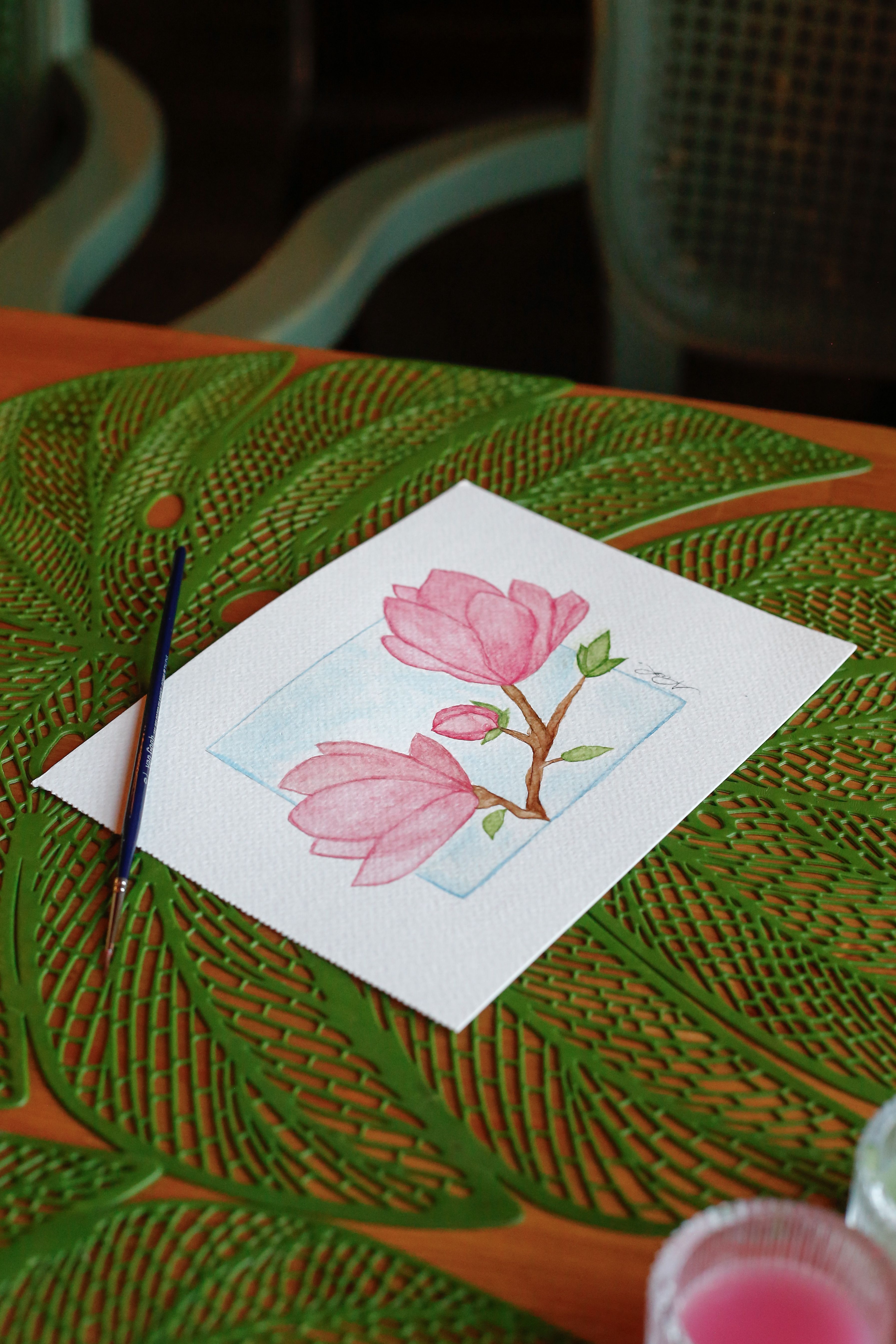 A beautiful watercolor painting of pink magnolia flowers on a leaf-patterned mat with a brush nearby.