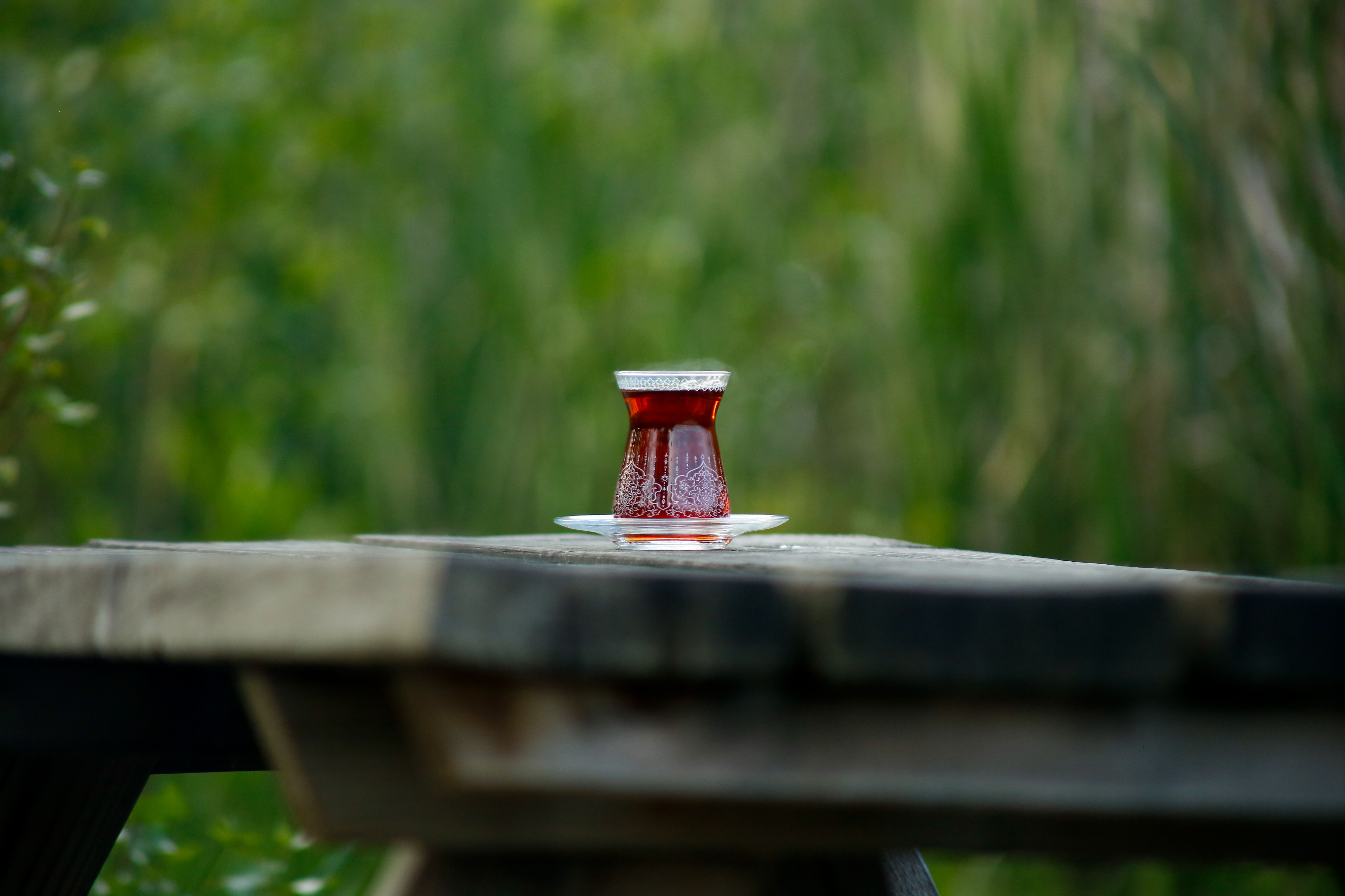 A traditional glass of Turkish tea placed on an outdoor wooden picnic table with a natural green background.