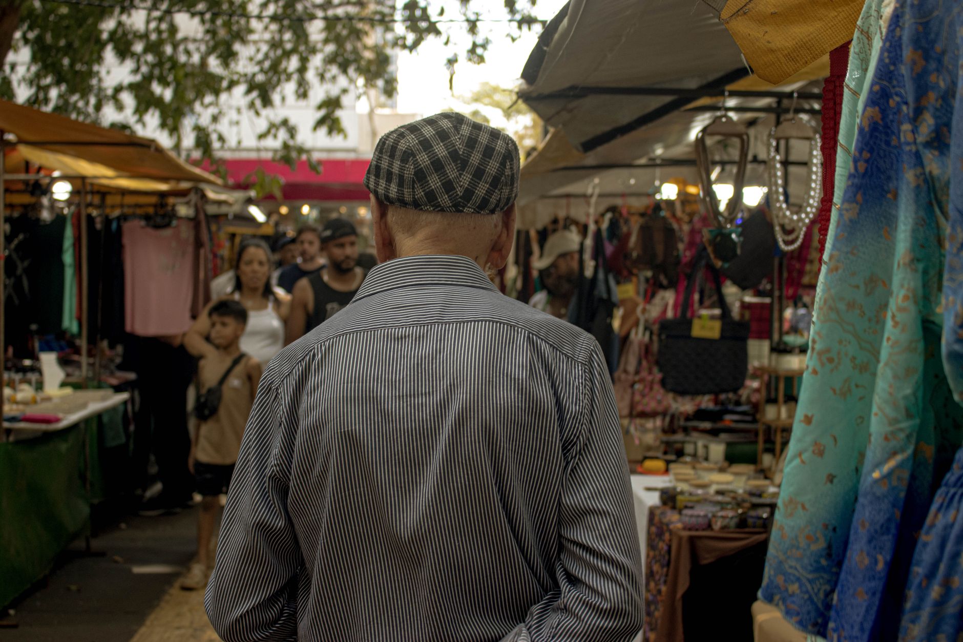 A senior adult in a cap explores a vibrant outdoor market, surrounded by stalls and people.