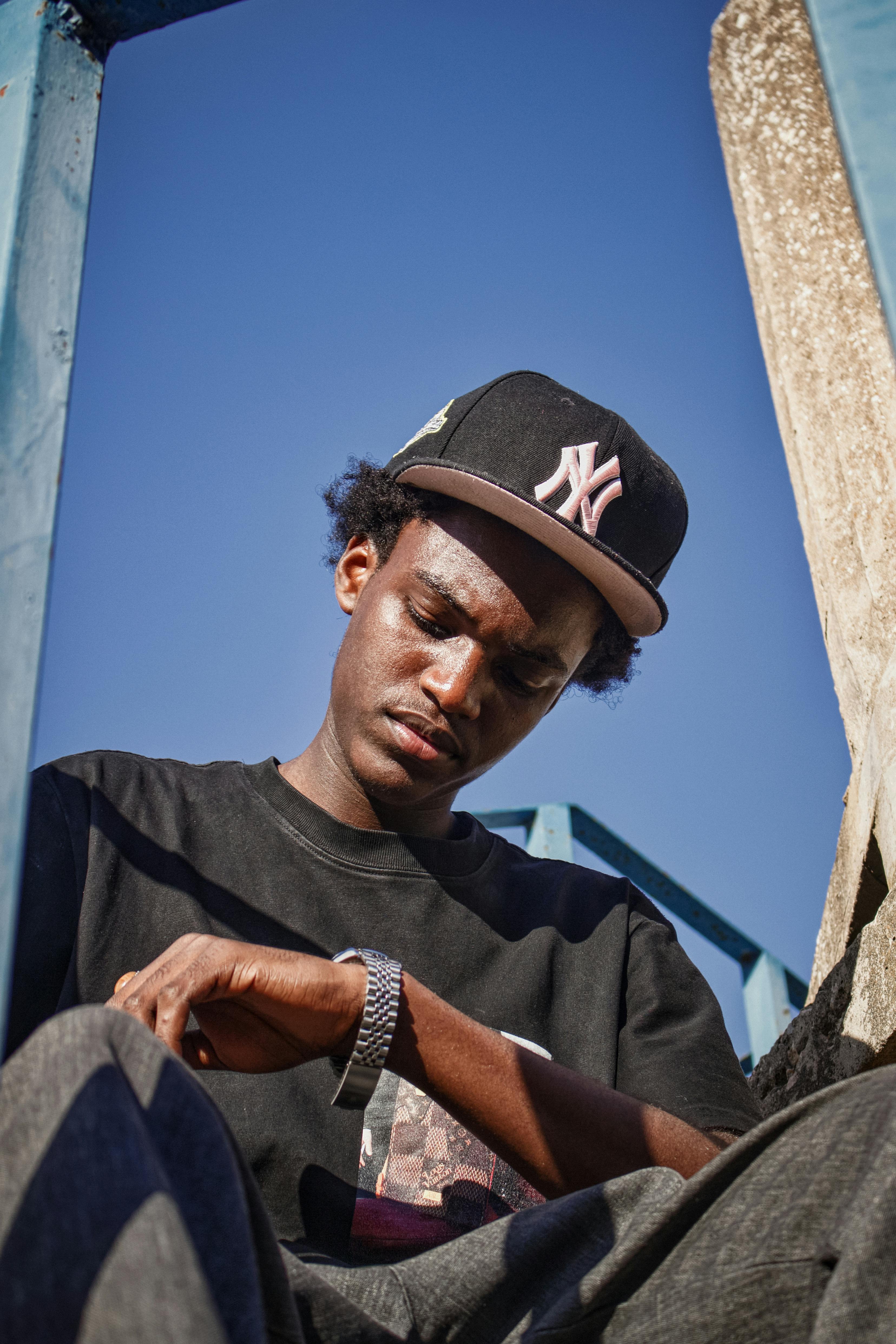 Man in casual clothes sitting outside, wearing a cap and watch, against a blue sky.