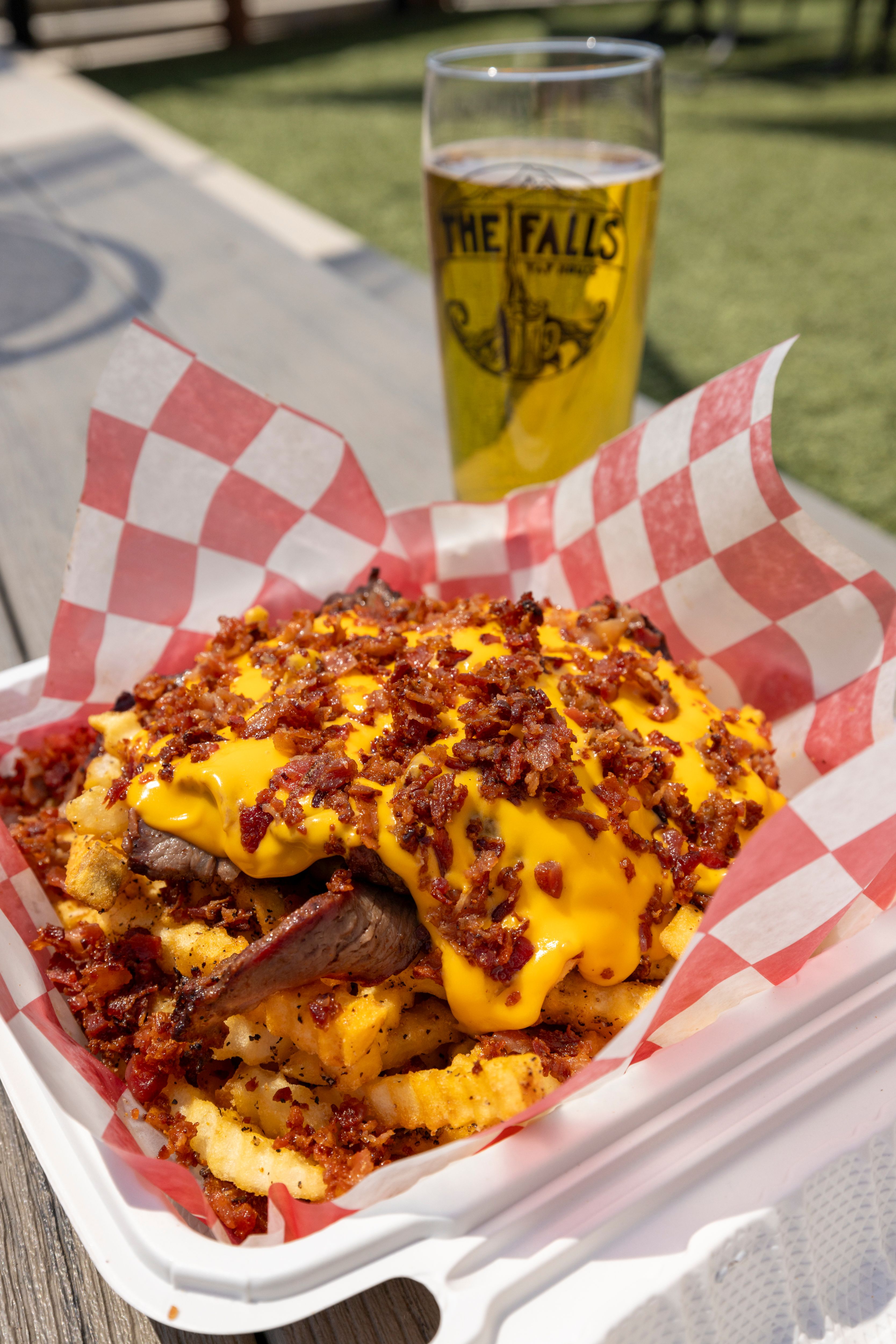 Nobleseed Nobleseed - Delicious loaded cheese fries with bacon on a picnic table, paired with a refreshing beer in a sunny outdoor setting.