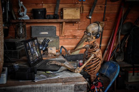 A skeleton with glasses and headphones using a laptop in a cluttered workshop setting.