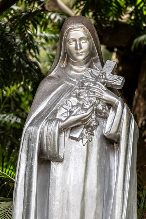 Free A silver statue of a saint holding a cross and roses, surrounded by lush greenery, radiating peace and reverence. Stock Photo