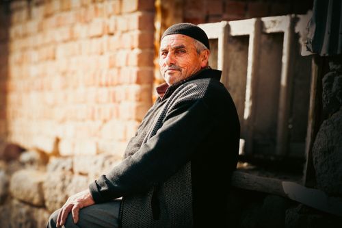 Free Portrait of an elderly man sitting outdoors against a sunlit brick wall in casual attire. Stock Photo